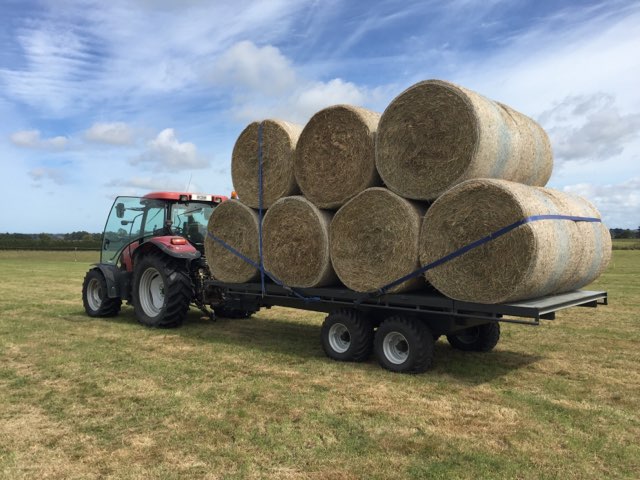 Maxi T100 with fold-down headboard and hay extension carrying strapped round bales behind a tractor