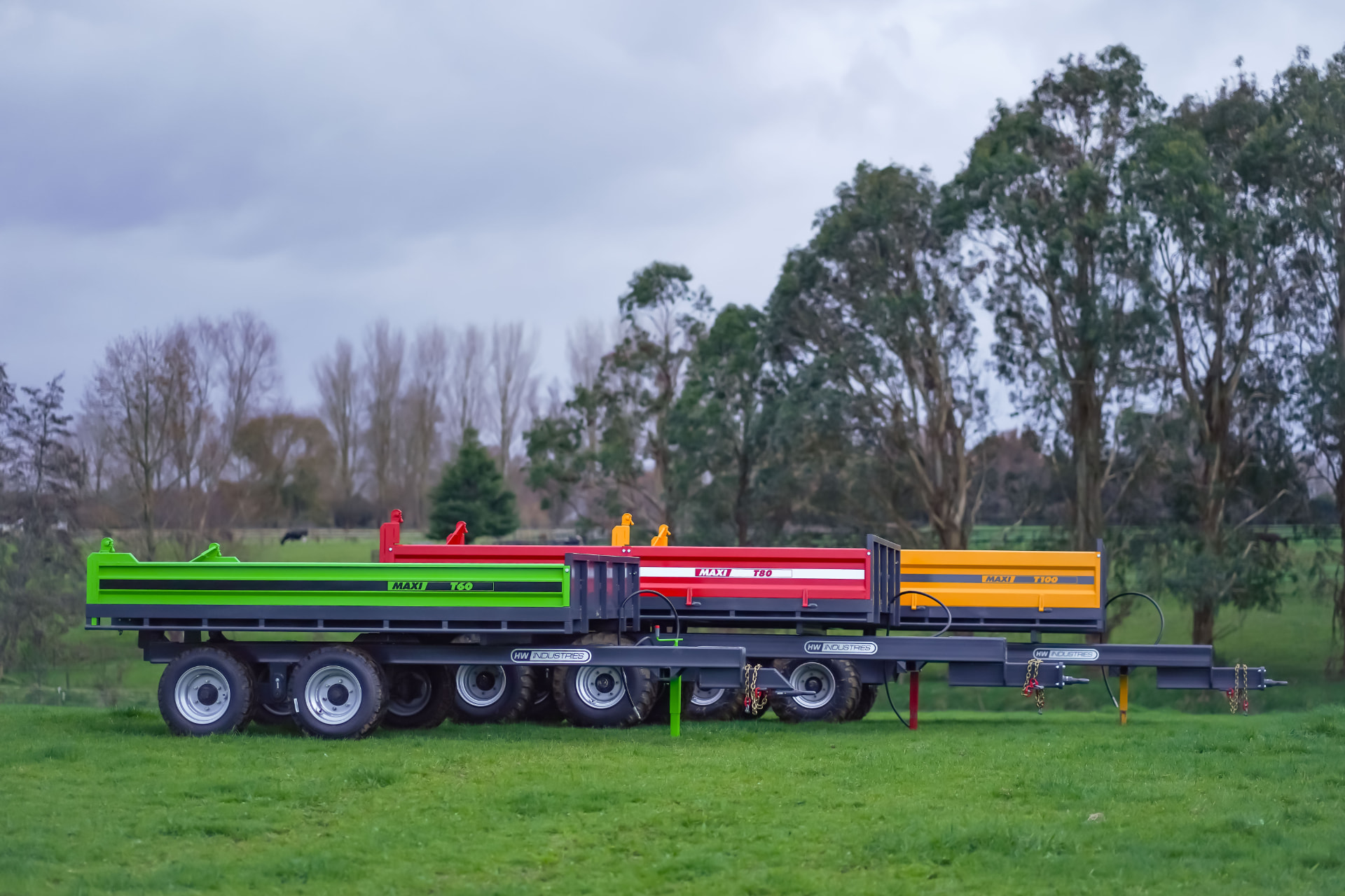 Green, red and orange Maxi tip trailers lined up on a grassy paddock