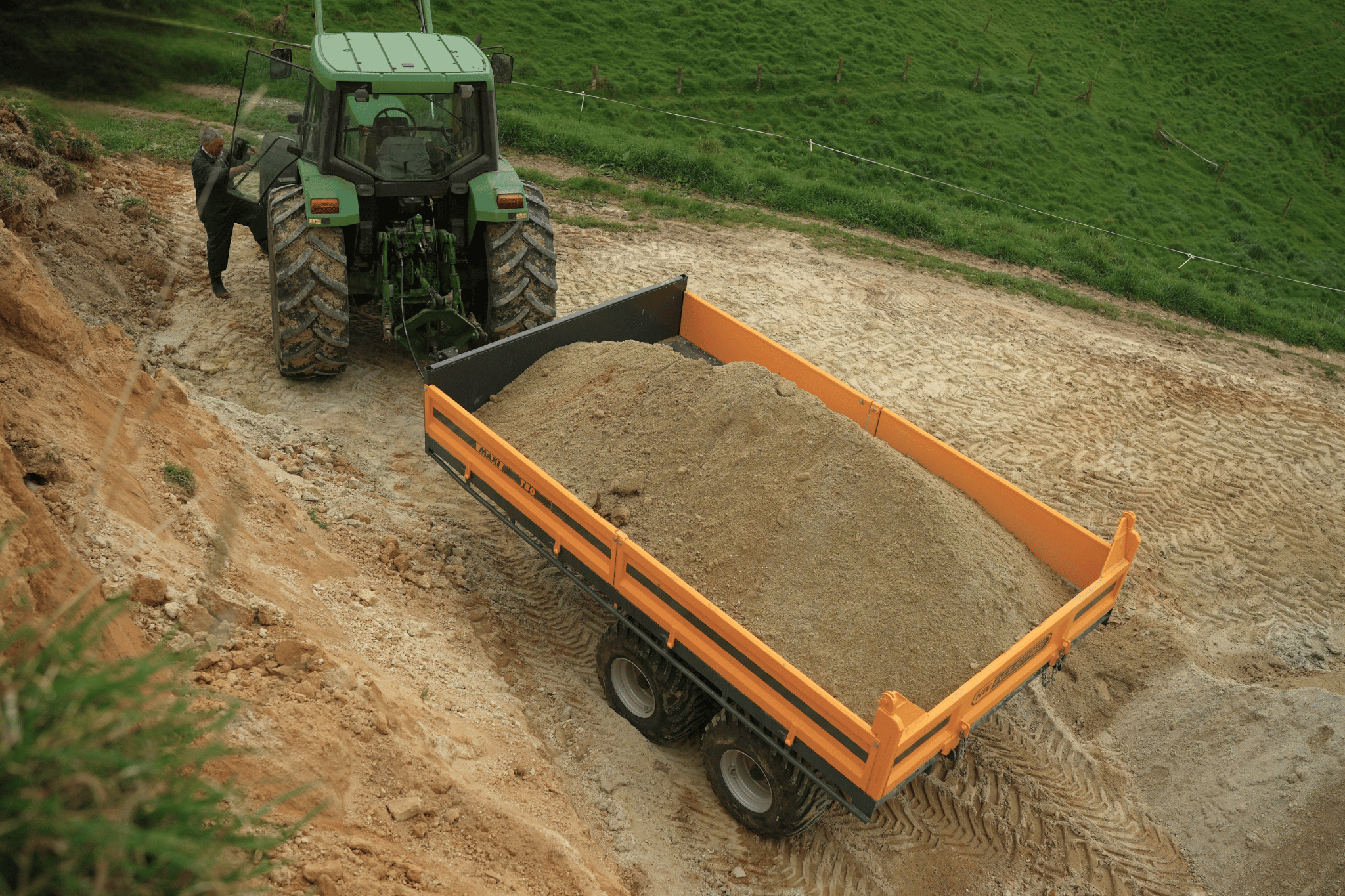 Loaded Maxi T80 tip trailer behind a John Deere tractor parked on a farm track cut into a hillside